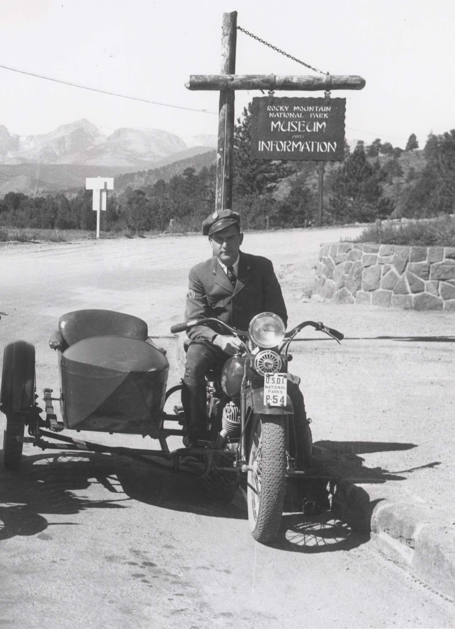 Man in NPS uniform sits astride a motorcycle with sidecar attached beneath a park sign. He wears a shield-shaped badge on his cap.